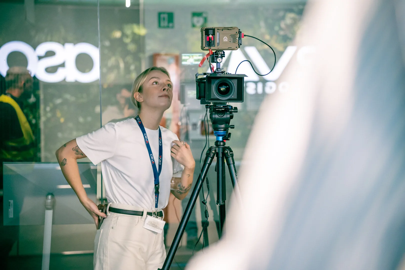 Malwina in a customer warehouse, looking up alongside a tripod-mounted cinema camera during a Nomagic video shoot.
